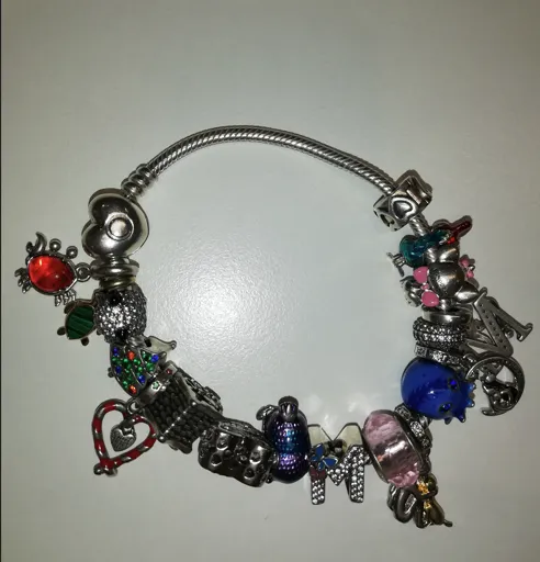 Eine silberne Charm-Armband mit verschiedenen Anhängern, darunter ein Herz, ein Haus, eine Maus und ein Buchstabe M. Das Armband hat eine Mischung aus bunten Perlen und Steinen.