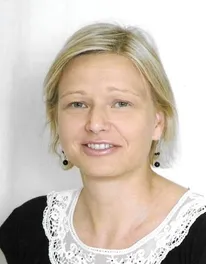 Eine Frau mit kurzen blonden Haaren, die ein schwarzes und weißes Oberteil und Ohrringe trägt. Sie lächelt leicht und sieht direkt in die Kamera.