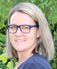 Eine Frau mit kurzen blonden Haaren, die eine blaue Brille und ein blaues Hemd trägt, lächelt draußen in der Nähe einiger grüner Blätter.