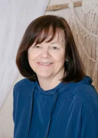 Eine Frau mit kurzen, braunen Haaren und einem warmen Lächeln trägt eine blaue Kapuzenjacke. Sie steht vor einer strukturierten weißen Wand.