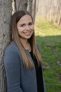 Eine Frau mit langen braunen Haaren lächelt in die Kamera. Sie trägt einen grauen Cardigan über einem schwarzen Oberteil und lehnt sich an einen Baumstamm. Der Hintergrund ist ein grasbewachsener Bereich mit Bäumen.