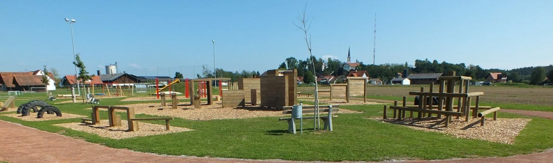 Ein Outdoor-Spielplatz mit Holzstrukturen, einem Rasenfeld und einem Mülleimer in der Nähe einer Bank. Eine Spielplatzrutsche befindet sich links.