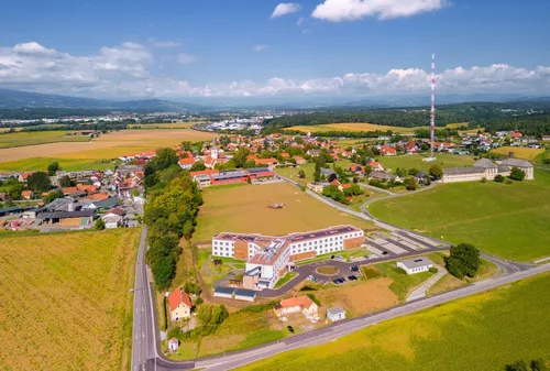 Luftaufnahme einer Stadt mit einem großen Gebäude, mehreren Häusern, grünen Feldern, einem Turm und einer Straße.