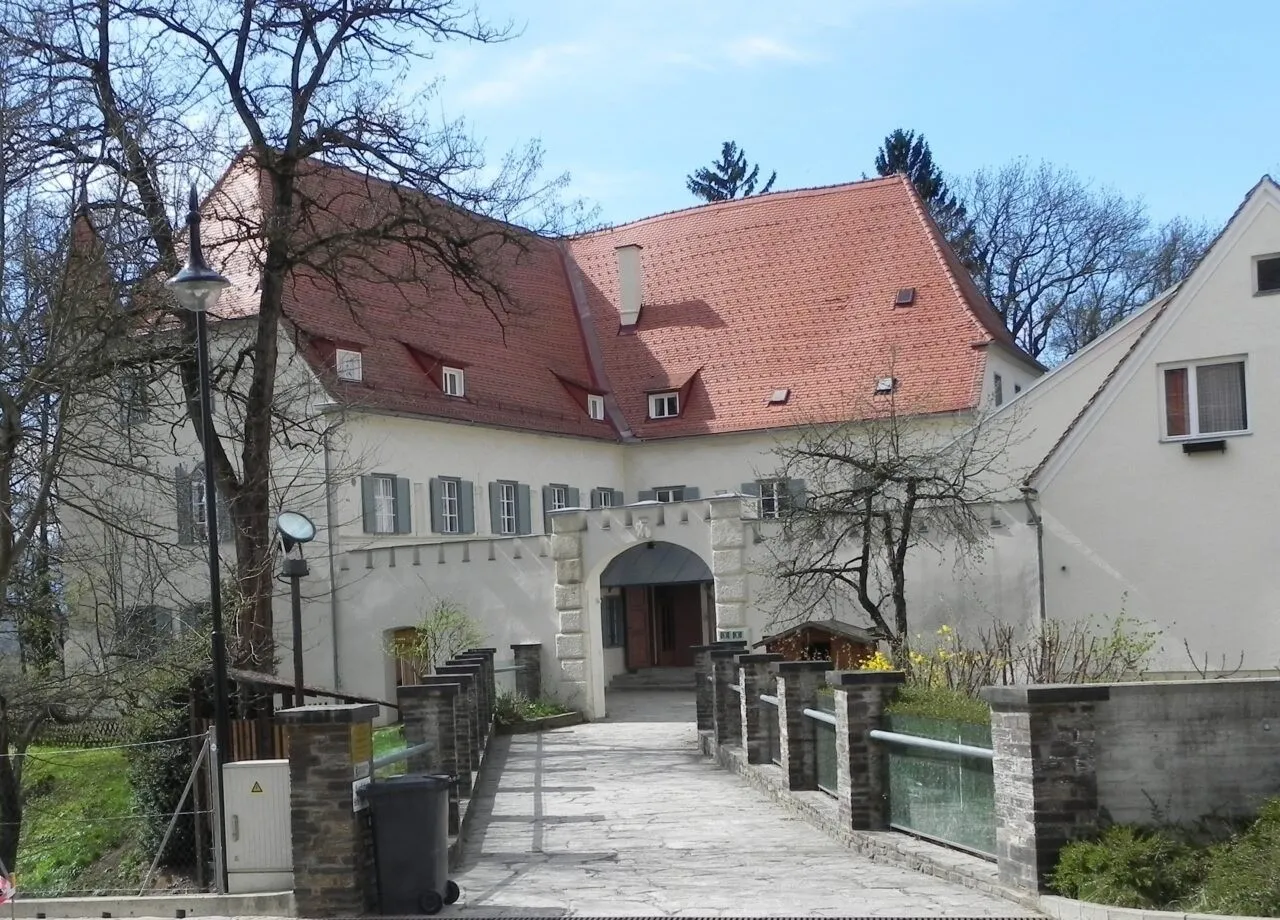 Ein großes weißes Gebäude mit einem roten Dach befindet sich an einem sonnigen Tag. Links steht ein Baum und eine Straßenlaterne. Ein Mülleimer steht neben dem Gehweg.