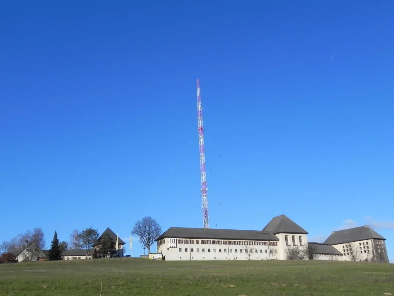 Ein großes weißes Gebäude mit vielen Fenstern steht auf einem grünen Hügel, mit einem hohen Turm in der Mitte und einem klaren blauen Himmel darüber.