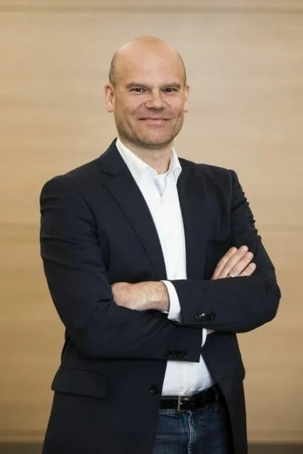 Ein kahlköpfiger Mann in einem dunklen Anzug mit verschränkten Armen, lächelt vor einer hellen Wand.