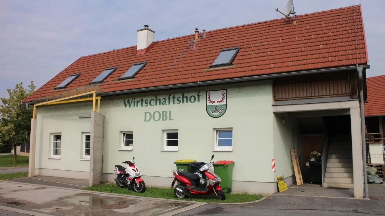 Ein Gebäude mit roten Ziegeln auf dem Dach, einem Logo über dem Eingang und zwei Rollern davor. Das Gebäude hat drei Fenster und einen Schornstein.