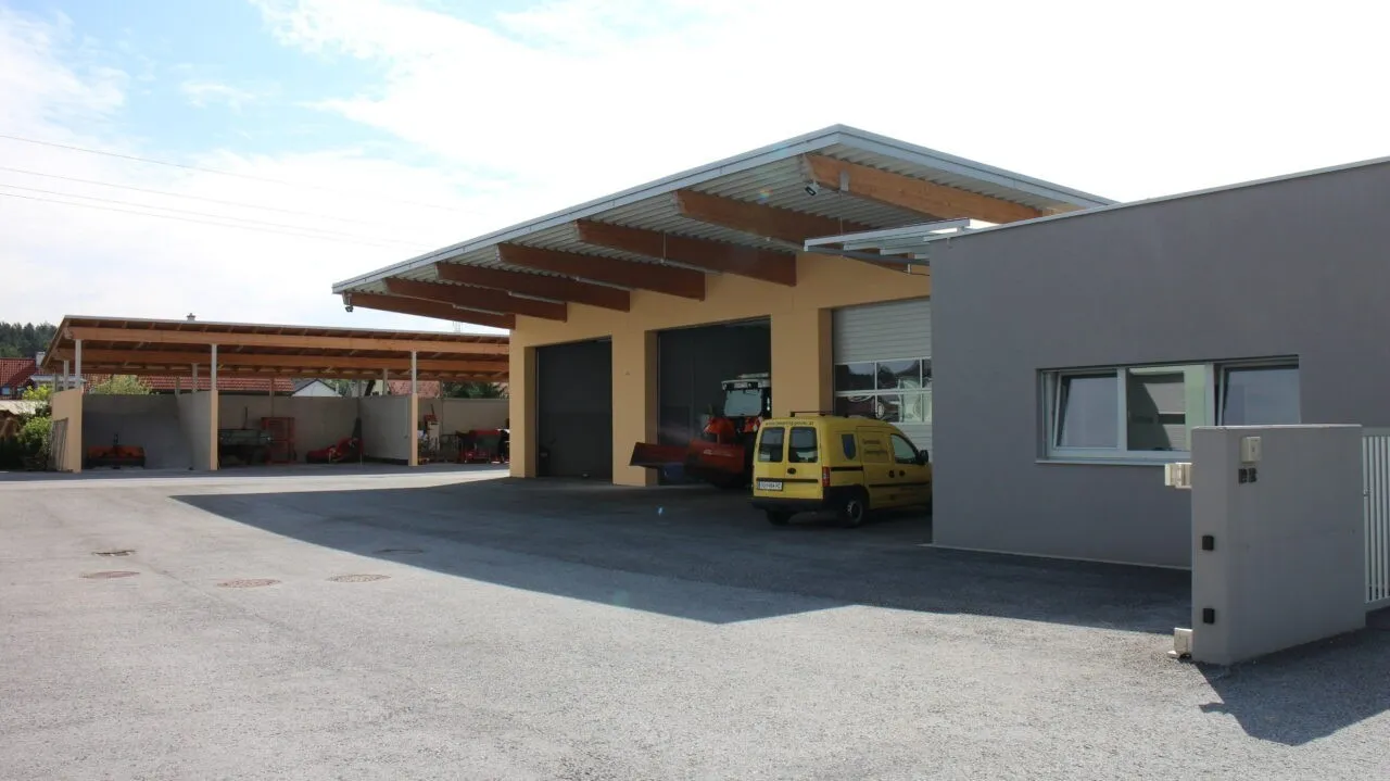 Ein gelber Lieferwagen steht vor einem gelben Gebäude mit offener Garage, einem roten Traktor im Inneren und Überwachungskameras.