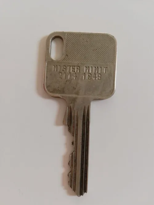 Ein Metallschlüssel mit der Aufschrift Mister Minit 2114 1048 auf weißem Hintergrund.