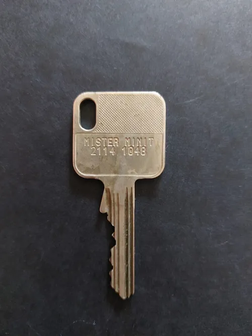Ein goldfarbener Schlüssel mit der Aufschrift 'Mister Minit 2114 1048' liegt auf einer schwarzen Oberfläche. Der Schlüssel hat eine strukturierte Oberseite und sichtbare Rillen.