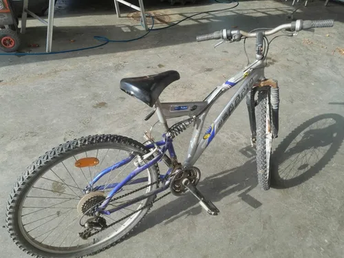 Ein blaues und silbernes Fahrrad mit schwarzem Sattel steht auf einer Betonfläche. Das Fahrrad hat einen Reflektor am Hinterrad und einen silbernen Rahmen.