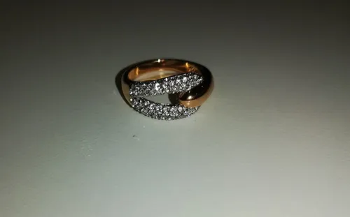 Ein goldener Ring mit zwei ineinander verschlungenen Bändern, verziert mit mehreren kleinen Diamanten, wird auf einer flachen, neutral gefärbten Oberfläche gezeigt.