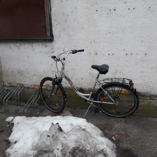 Ein silbernes Fahrrad mit schwarzem Sattel steht auf dem Bürgersteig, mit Schnee am Boden und einem Fahrradständer in der Nähe.