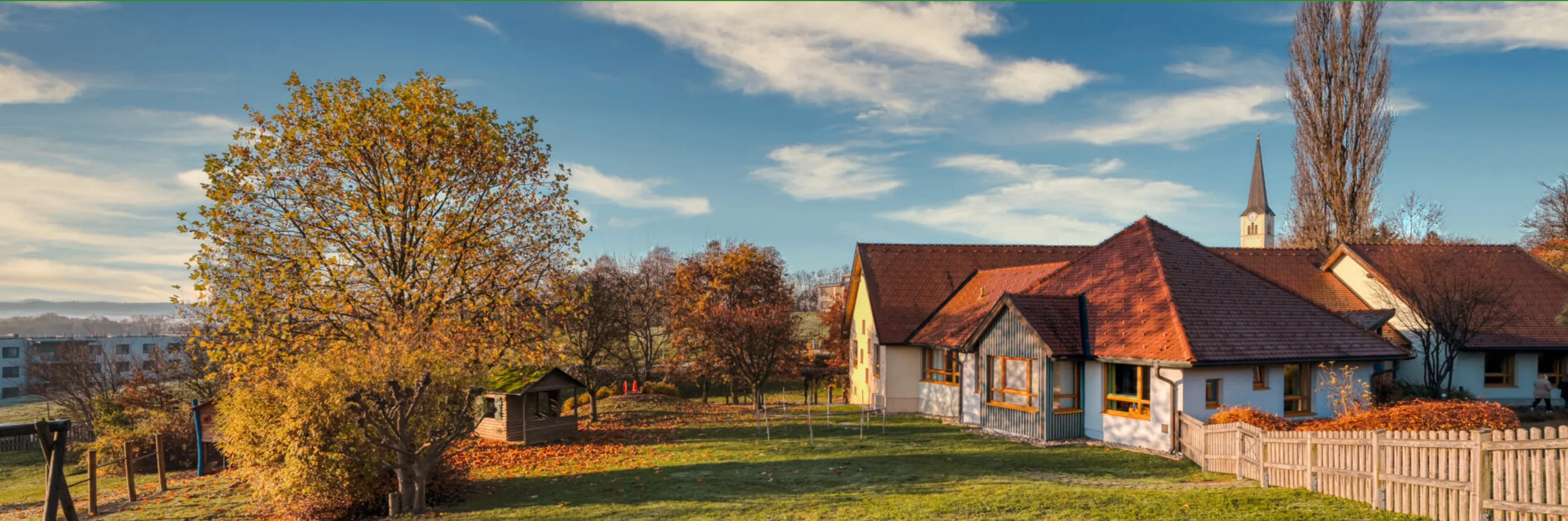 Ein Haus mit einem braunen Dach und gelben Fenstern steht auf einem Rasenfeld mit Herbstbäumen und einem bewölkten Himmel.