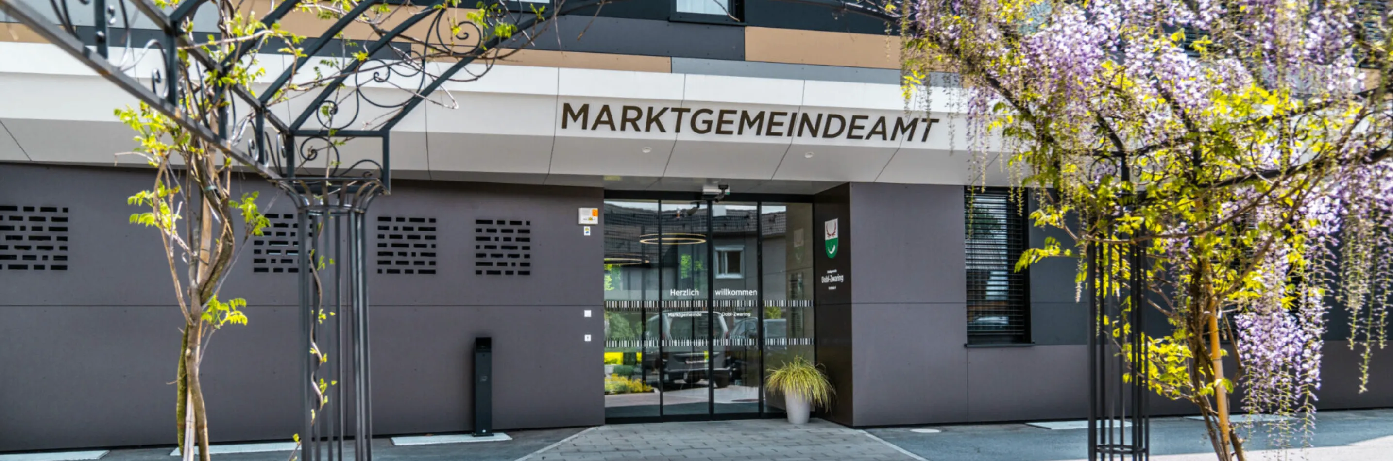 Der Eingang eines Gebäudes mit der Aufschrift 'MARKTGEMEINDEAM'. Es verfügt über eine Glastür, eine Überwachungskamera und einen Topf mit einer Pflanze. Auf der rechten Seite des Eingangs befindet sich ein Logo.