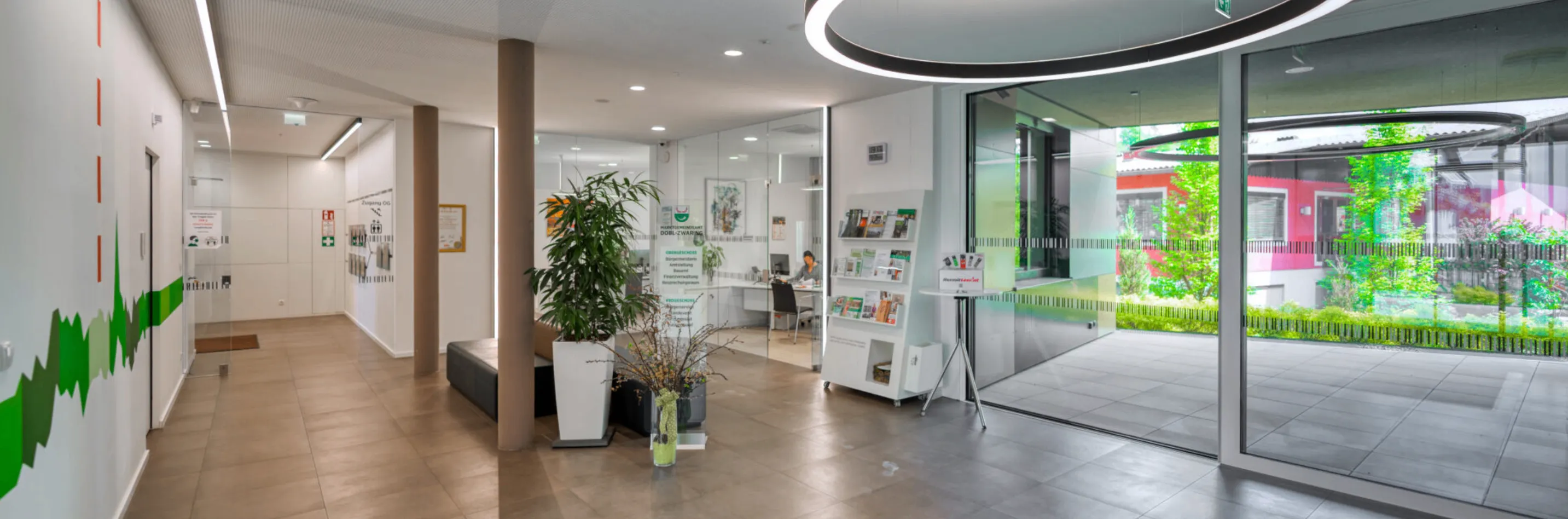 Ein Büro mit einer großen Topfpflanze und einer Glaswand. Es gibt einen Tisch mit Büchern darauf und eine Frau sitzt an einem Schreibtisch. Der Raum hat eine Glastür und eine kreisförmige Lichtinstallation an der Decke.