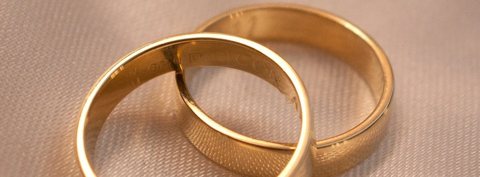 Zwei goldene Eheringe liegen auf einer weißen Oberfläche. Ein Ring hat 'LIEBE' eingraviert, der andere 'LÄCHELN'. Sie liegen eng beieinander.