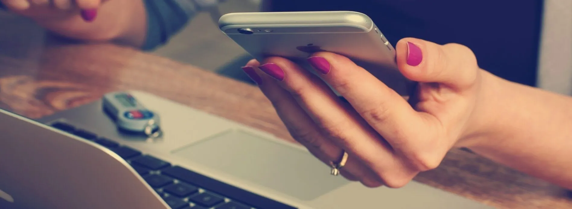 Eine Hand mit rosa Fingernägeln hält ein silbernes iPhone vor einer Laptop-Tastatur.