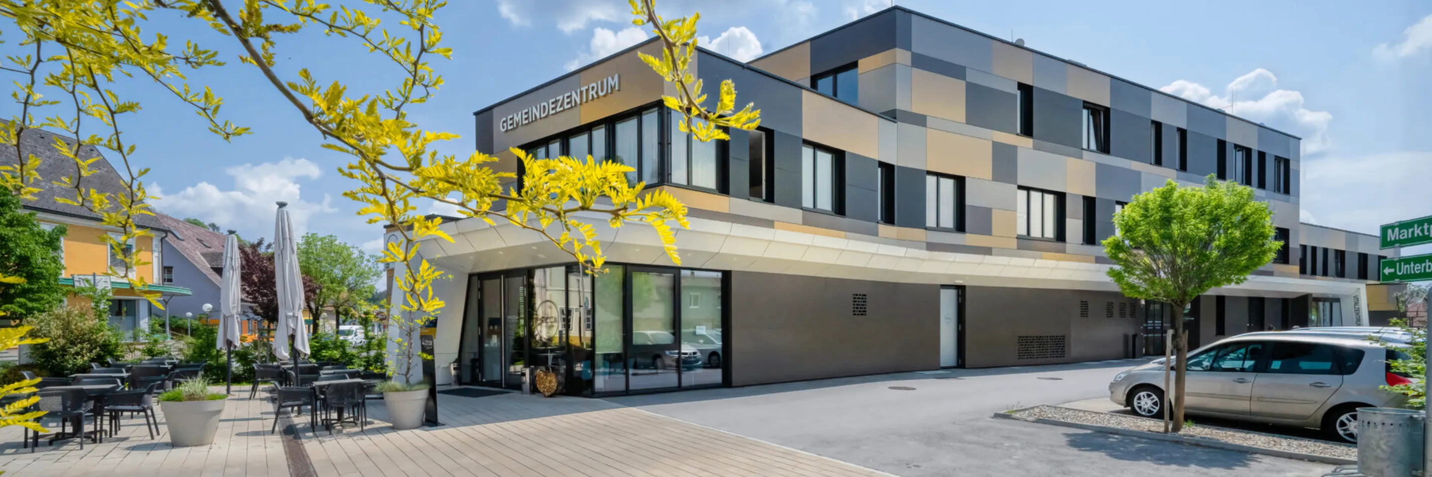 Die Vorderseite des Gemeindezentrum-Gebäudes mit gelben Blättern an einem Ast, Glastüren und geparkten Autos, die sich in den Fenstern spiegeln.