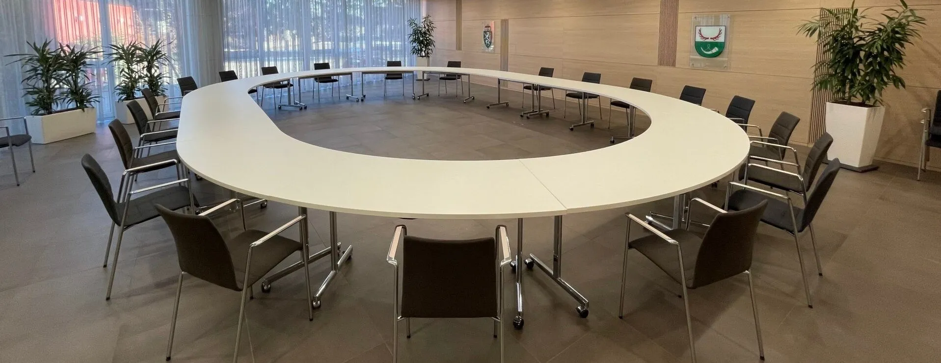 Ein großer, leerer Konferenzraum mit einem U-förmigen Tisch und Stühlen, einem Topfpflanzen und einer Wand mit einem Glasfenster.