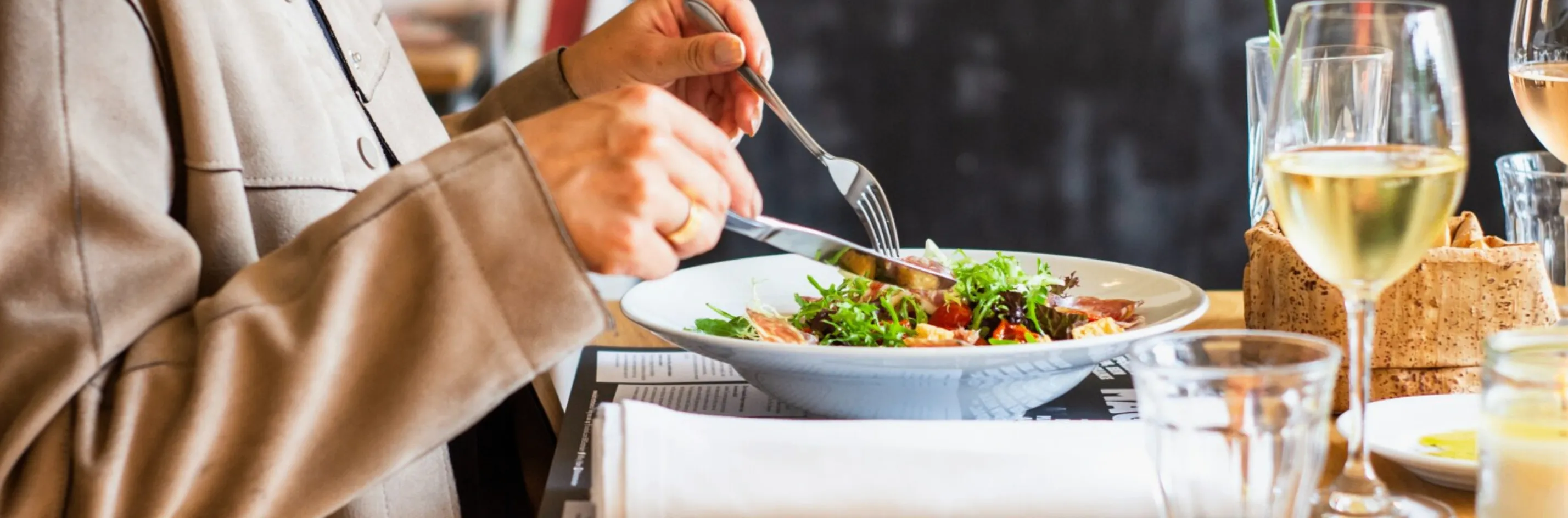 Eine Person mit Ring schneidet einen Salat in einer weißen Schüssel mit Messer und Gabel.