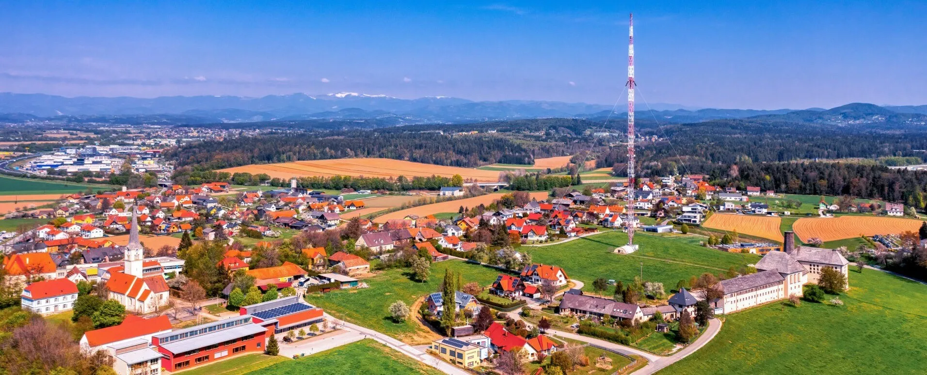 Luftaufnahme eines Dorfes mit roten Dächern, Solarpanelen und einem Turm, umgeben von Feldern, Bäumen und Bergen in der Ferne.