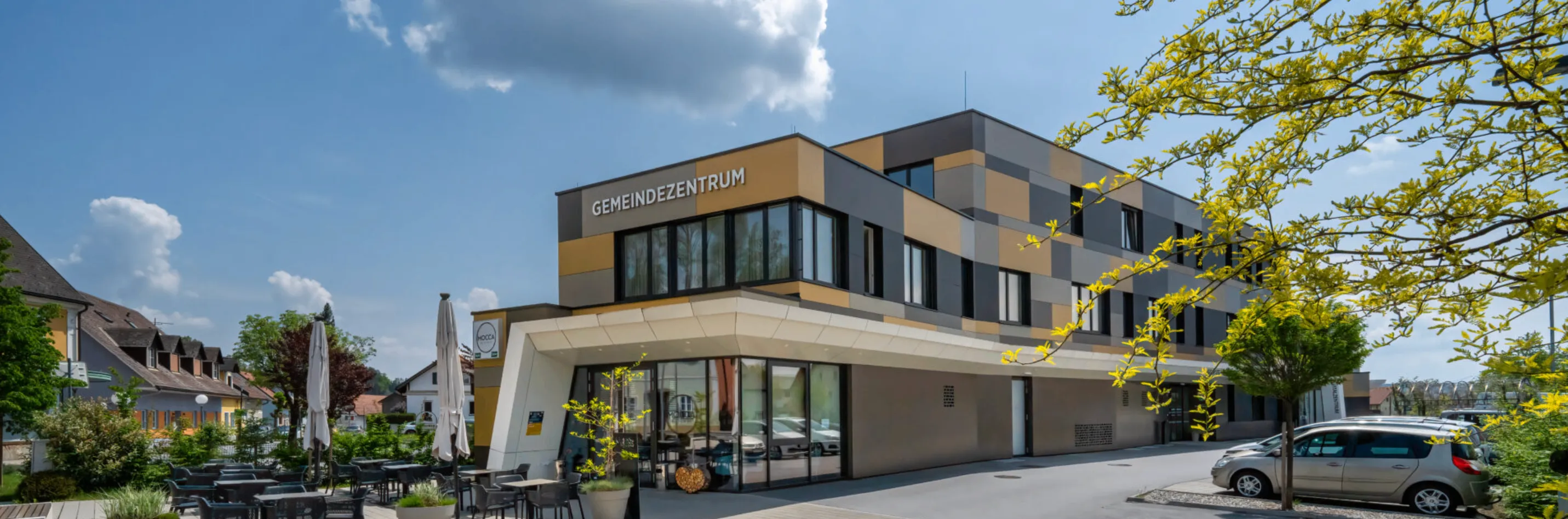 Ein modernes gelbes und braunes Gebäude mit dem Wort Gemeindezentrum oben. Es hat Glasfenster und Türen. Vor dem Gebäude steht eine Pflanze in einem Topf.