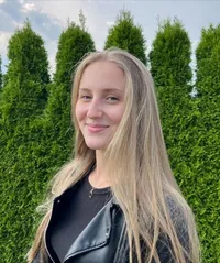 Eine Frau mit langen blonden Haaren lächelt vor einer Hecke. Sie trägt eine schwarze Lederjacke und ein schwarzes Shirt. Der Himmel ist blau mit einigen Wolken.
