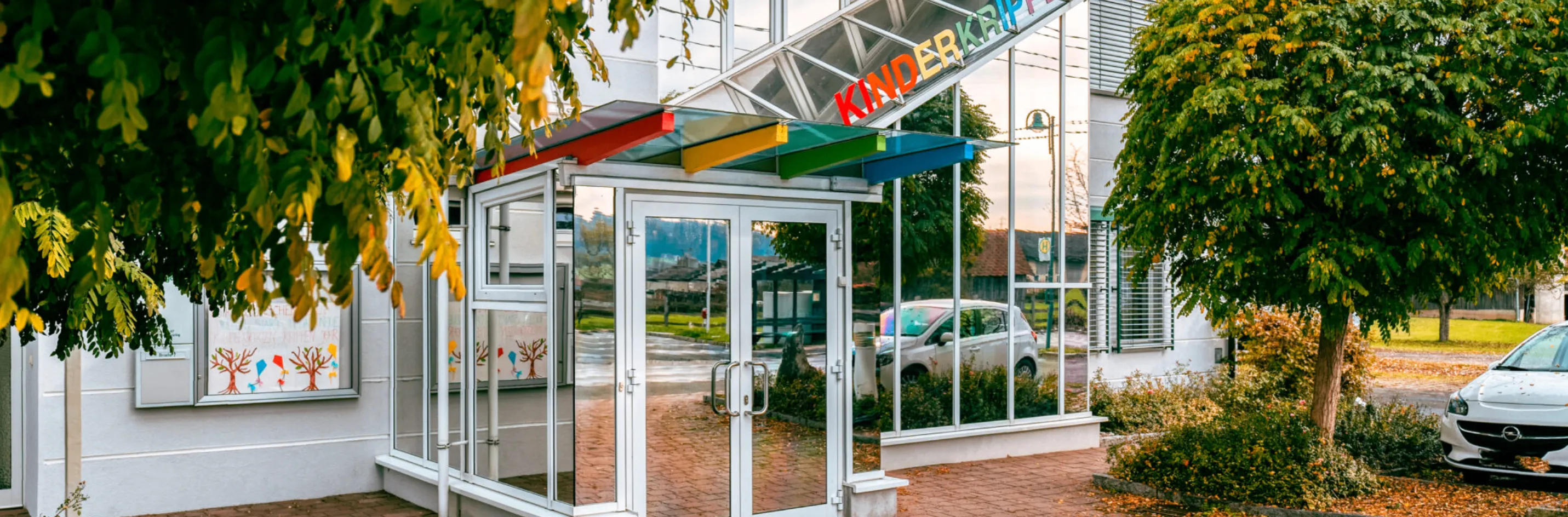 Ein glasverkleideter Eingang mit farbigem Rand, beschriftet mit 'KINDERKRAFT', verfügt über einen Ziegelweg und reflektierende Glaswände. Ein Auto ist draußen sichtbar.