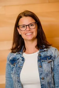 Eine Frau mit braunen Haaren lächelt und trägt eine Jeansjacke und eine Halskette. Sie hat eine Brille und ein weißes Hemd. Der Hintergrund ist hölzern.
