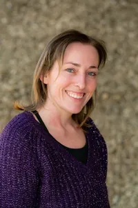 Eine Frau mit blonden Haaren lächelt in die Kamera. Sie trägt einen lila gestrickten Pullover und steht vor einer Steinmauer.
