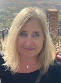 Eine lächelnde blonde Frau steht vor einem Bergpanorama und trägt ein schwarzes Shirt und eine Kreuzkette.
