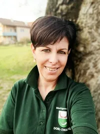 Eine Frau in einem grünen Poloshirt mit dem Wort DOBL-ZWANG steht neben einem Baum und lächelt.