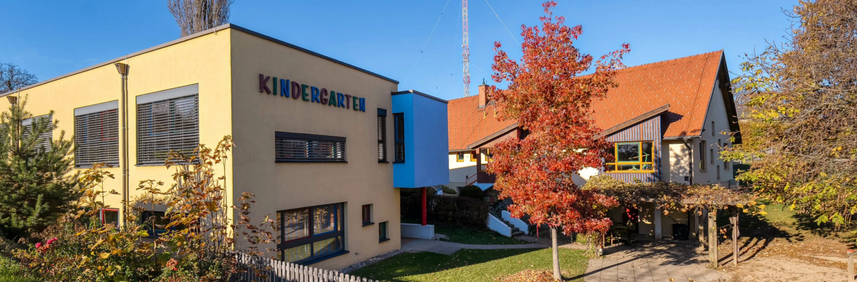 Ein Kindergartengebäude mit einer blauen Erweiterung steht vor einem Haus. Ein Herbstbaum mit roten Blättern steht im Vordergrund. Ein hoher Turm ist im Hintergrund zu sehen.