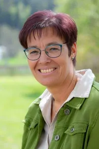 Eine Frau mit kurzen Haaren und Brille, lächelt und steht auf einem grünen Feld. Sie trägt eine grüne Jacke mit weißen Akzenten und eine Halskette.