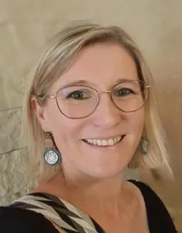 Eine Frau mit blondem Haar, die eine Brille und ein schwarzes Oberteil mit einem silbernen Ohrring trägt, lächelt in die Kamera.