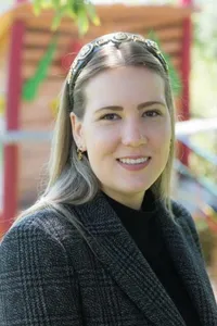 Eine Frau mit blonden Haaren lächelt, trägt ein schwarzes Rollkragenshirt und eine graue Jacke und eine Stirnband. Sie steht draußen in der Nähe eines Spielplatzes.
