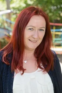 Eine Frau mit langen roten Haaren lächelt vor einem farbenfrohen Spielplatz. Sie trägt ein weißes Oberteil und eine blaue Strickjacke.