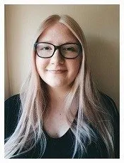 Eine Frau mit langen blonden Haaren und Brille in einem schwarzen Shirt posiert für ein Foto vor einem schlichten beigefarbenen Hintergrund.