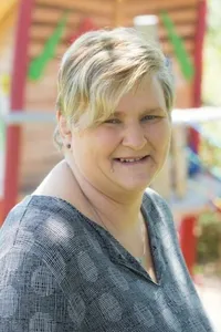 Eine Frau mit kurzen blonden Haaren lächelt in die Kamera, trägt ein graues Oberteil und steht vor einem Kinderspielplatz.