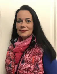 Eine Frau mit langen dunklen Haaren steht vor einer weißen Wand und trägt eine rot-schwarz gemusterte Weste und einen rosa Schal. Sie lächelt.