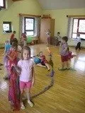 In einem Raum mit Holzböden beschäftigen sich mehrere junge Kinder mit verschiedenen Aktivitäten, einschließlich des Spielens mit einem Hula Hoop und schwebenden Luftballons. Die Kinder sind in Freizeitkleidung gekleidet.