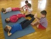 Fünf Kinder üben Yoga auf blauen Matten, liegend und dehnend in einem Raum mit Holzböden. Zwei Kinder interagieren, indem sie sich an den Händen halten.