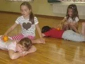 Vier junge Mädchen sind auf einem Holzboden und spielen. Zwei liegen auf dem Boden, eine mit einer Orange, die andere mit einem Spielzeug. Ein weiteres Mädchen sitzt mit einer Orange, und ein viertes Mädchen sitzt hinter ihnen.