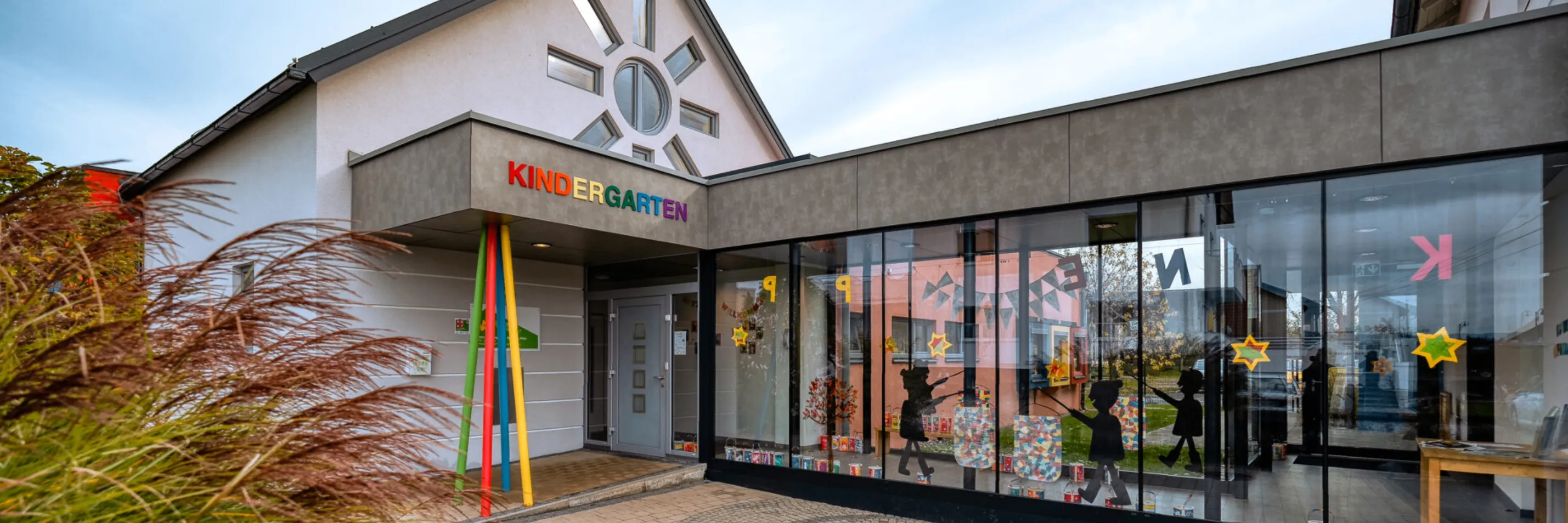 Die Vorderseite eines Kindergartengebäudes mit dem Wort Kindergarten in verschiedenen Farben geschrieben. Der Eingang hat eine Tür mit Glasscheiben und einem gelben Pfosten. Die Glasfenster zeigen eine Silhouette eines Kindes, das einen Beutel hält.