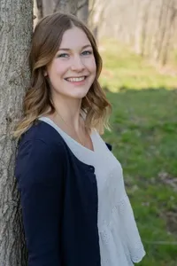 Eine lächelnde Frau mit blonden Haaren steht an einen Baumstamm gelehnt, trägt ein weißes Oberteil und eine marineblaue Strickjacke.