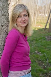 Eine blonde Frau mit langen Haaren, die ein rosa Sweatshirt trägt, lehnt sich in einem Grasbereich mit Bäumen im Hintergrund an einen Baum.