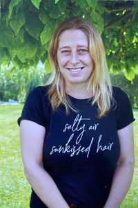 Eine Frau mit blonden Haaren, die ein schwarzes T-Shirt mit weißem Text trägt, lächelt unter einem Baum.