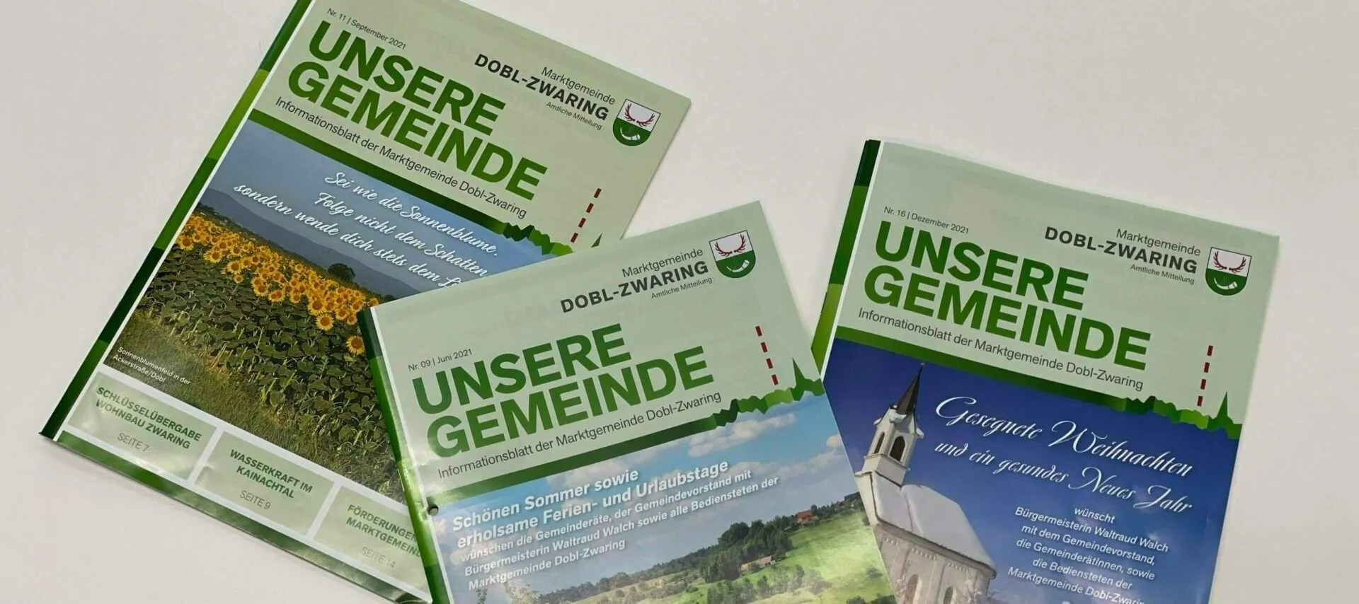 Drei grüne Broschüren mit den Titeln 'Unsere Gemeinde' und 'DOB-ZWARING' sind auf einer weißen Oberfläche zu sehen. Die erste Broschüre zeigt ein Landschaftsfoto und die zweite eine Kirche mit einem Turm.