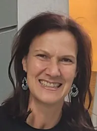Eine lächelnde Frau mit kurzen, gewellten braunen Haaren, die silberne Ohrringe und ein schwarzes Oberteil trägt.
